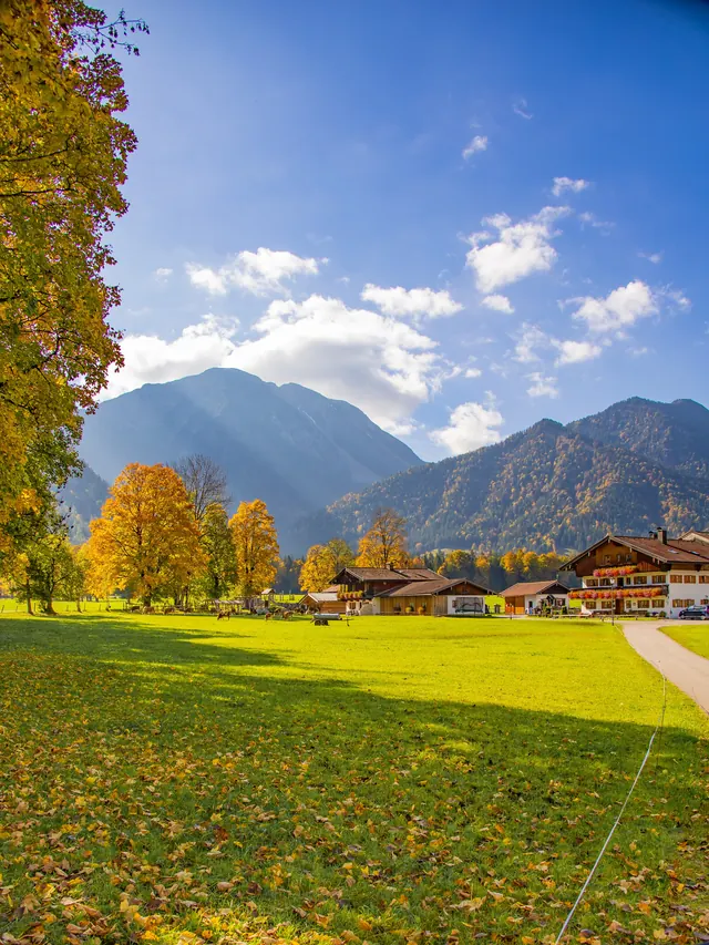 Herbstlich gefärbte Bäume vor Bergen, Häuser am Rand einer Wiese